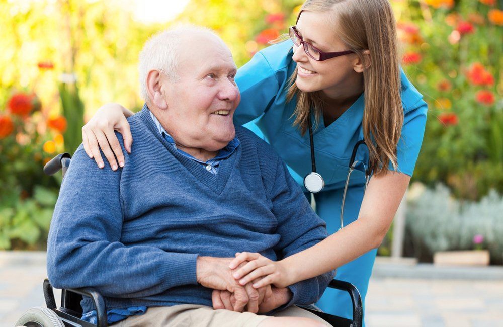 Young caregiver with stethoscope smiles at older man seated in wheelchair. They are outdoors, holding hands.