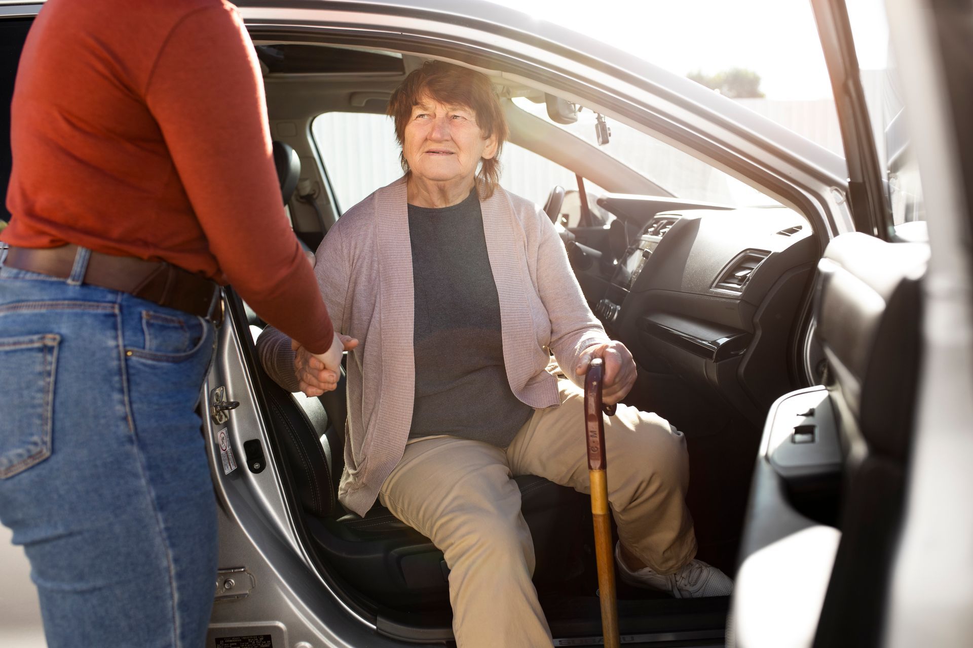 Elderly woman with a cane being helped out of a car by a caregiver.