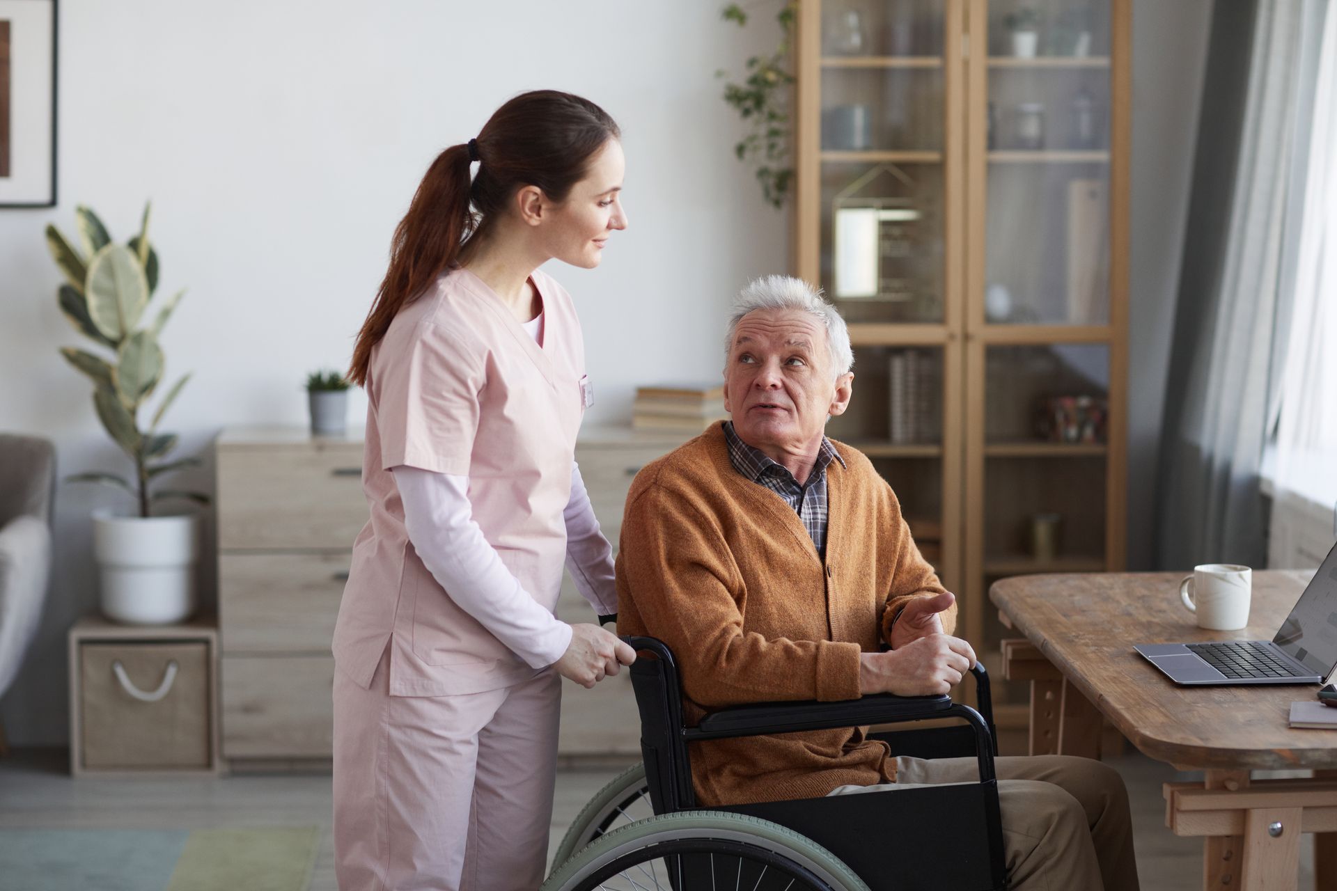 Caregiver assisting an older person in a wheelchair indoors near a table with a laptop.