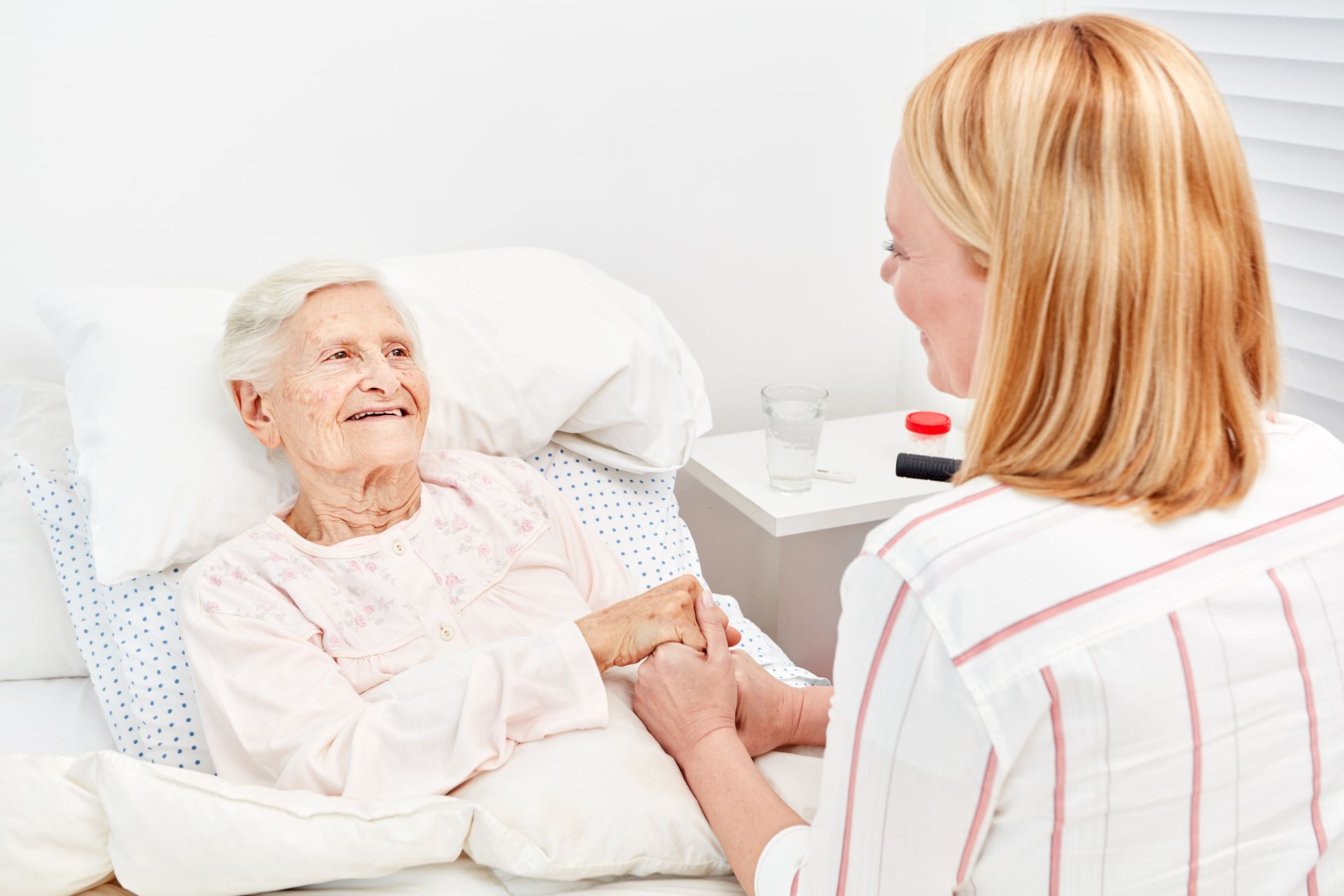 Woman in striped shirt holding hands with older person in bed, smiling.