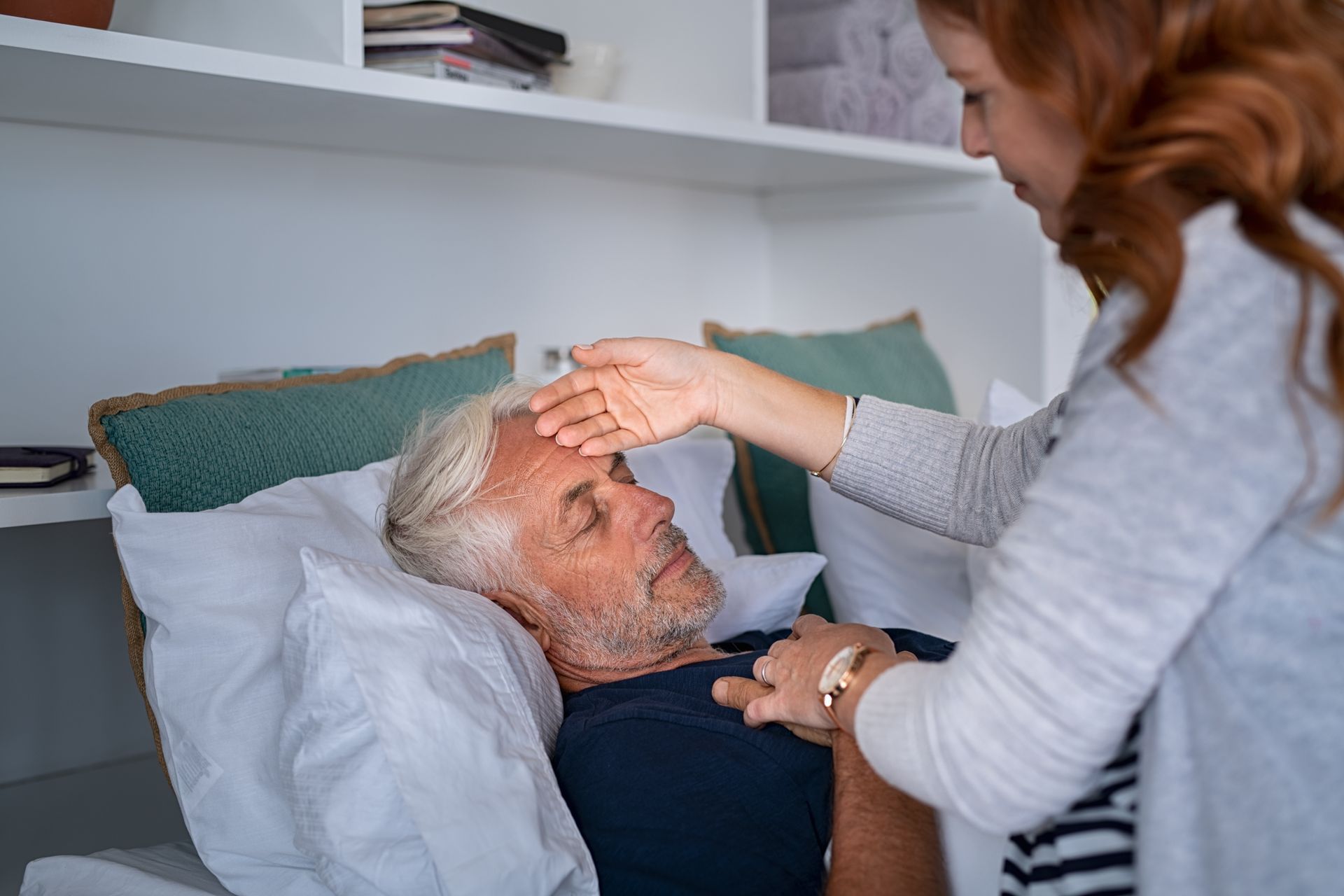 Woman checks the forehead of a person lying in bed; indoors, assessing health.
