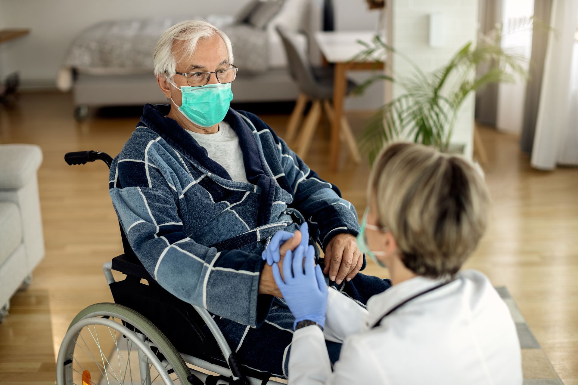 Elderly person in wheelchair wearing a face mask, receiving care from a healthcare worker, in a home.