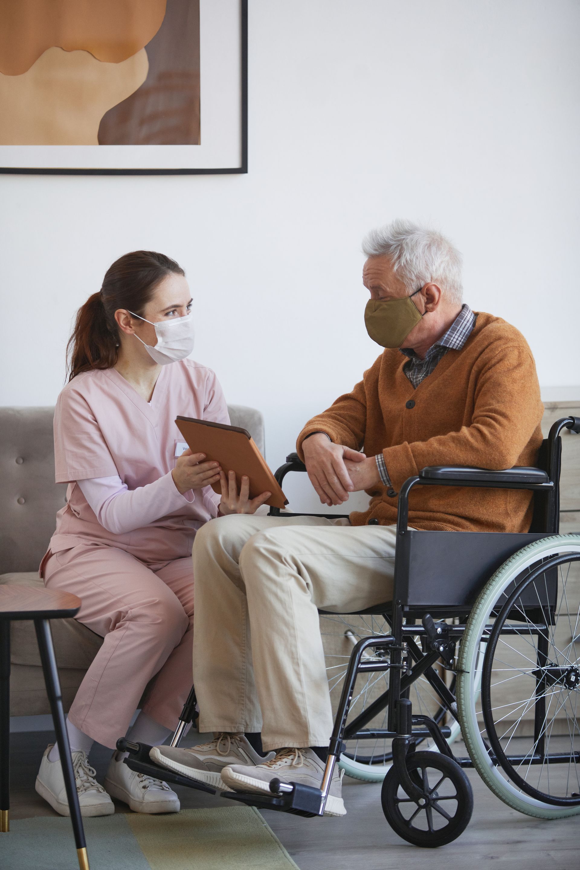 Nurse in pink scrubs and mask shows tablet to elderly person in wheelchair, both masked.
