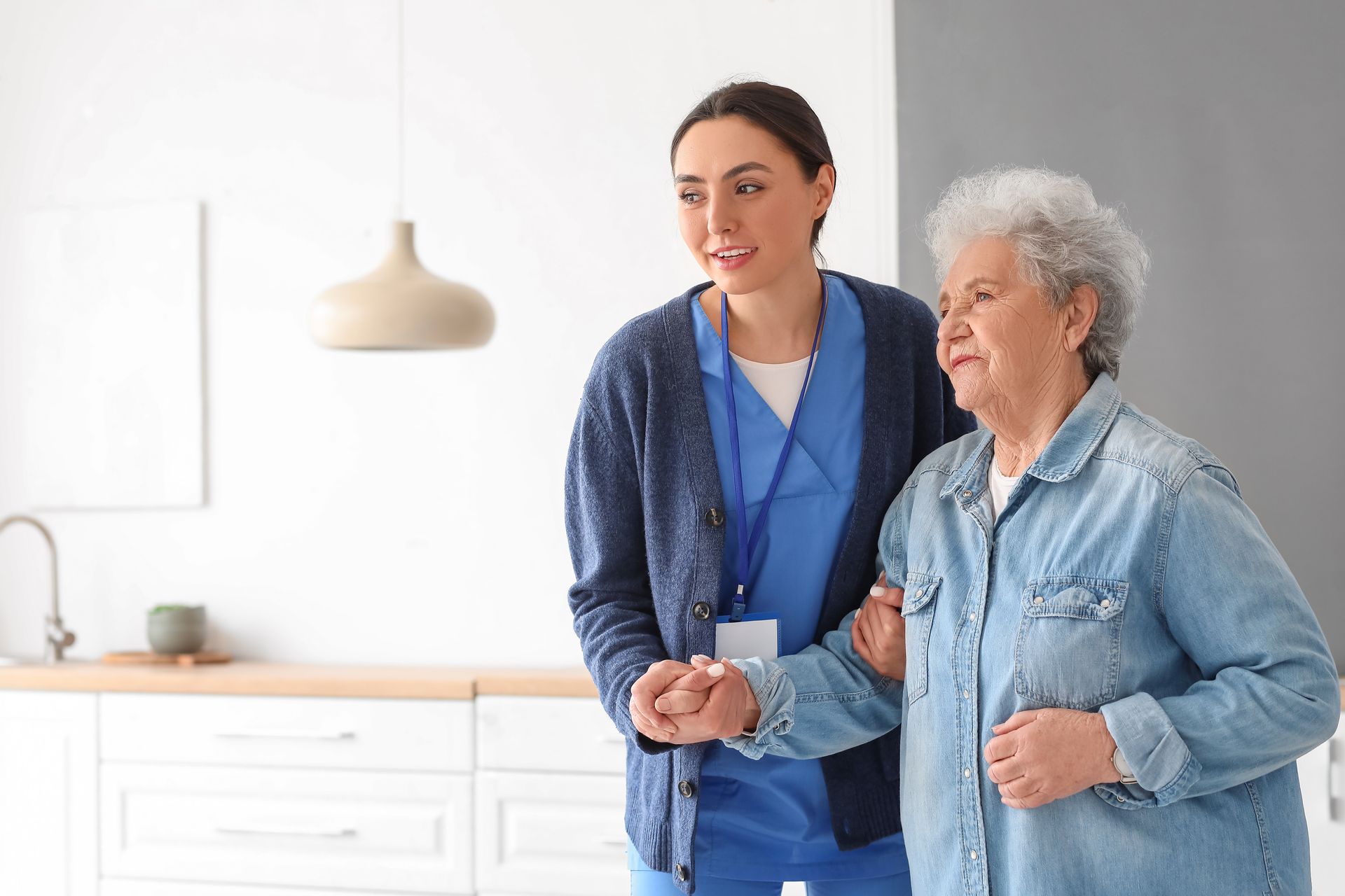 A caregiver assists an elderly woman walking indoors; both are smiling.
