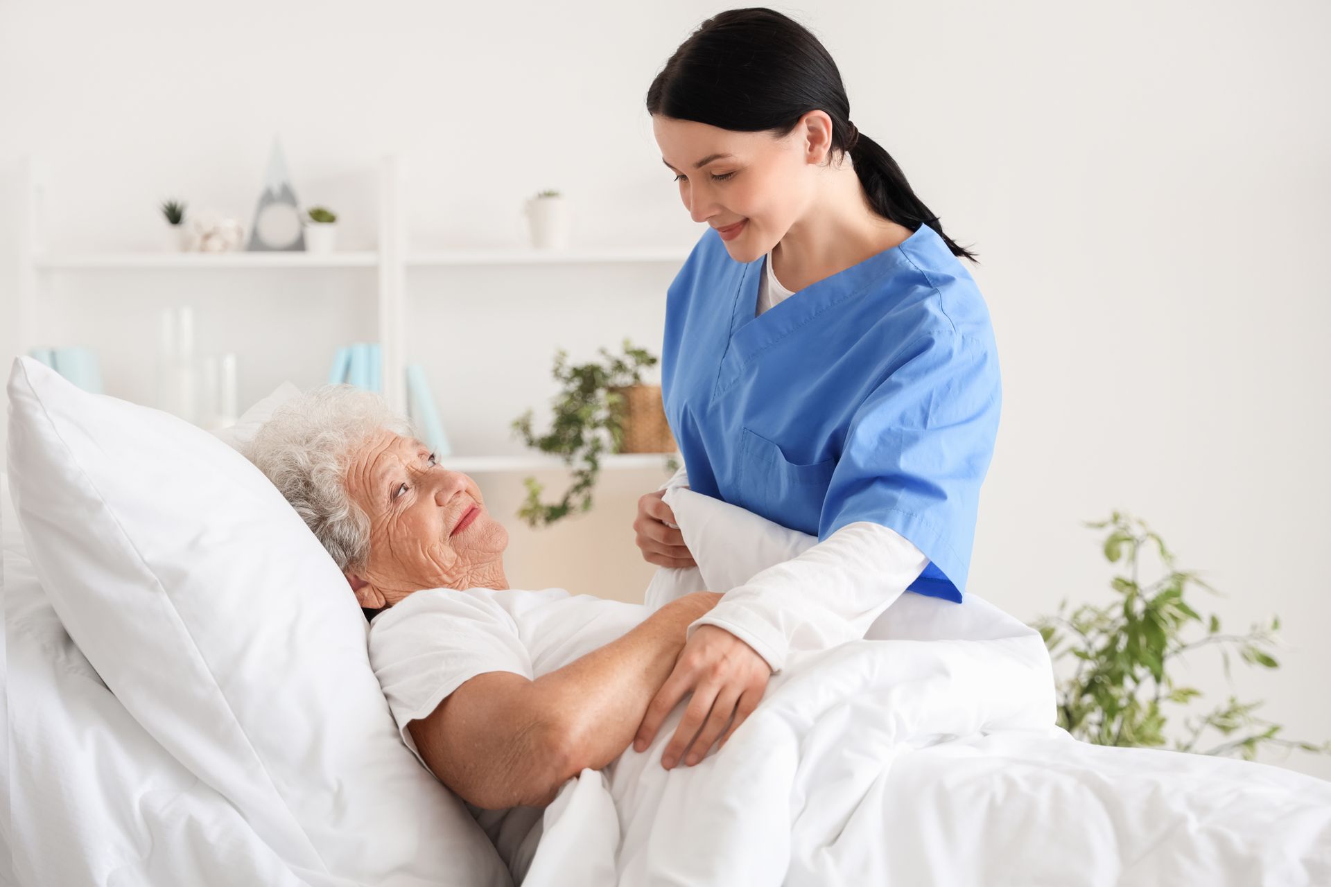 Nurse in blue scrubs attends to elderly patient in a hospital bed; white room, plants.