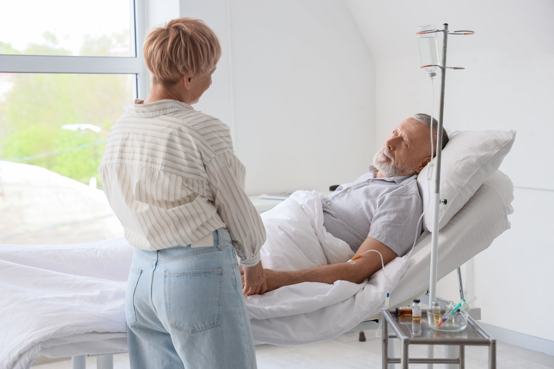Woman holding hands with a person in a hospital bed with an IV drip. Bright room, supportive gesture.