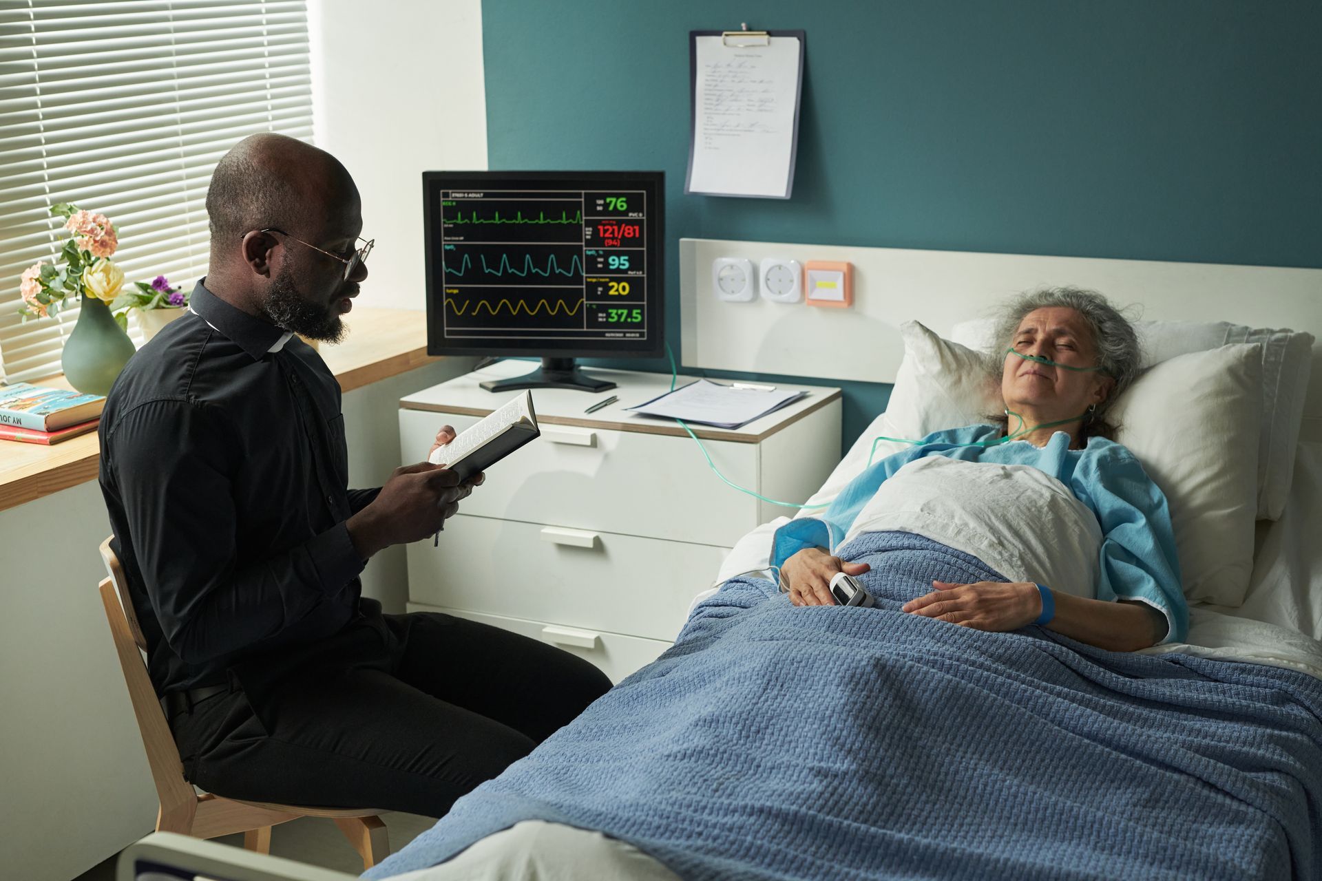 Man in clergy attire reads to person in hospital bed, monitor visible.