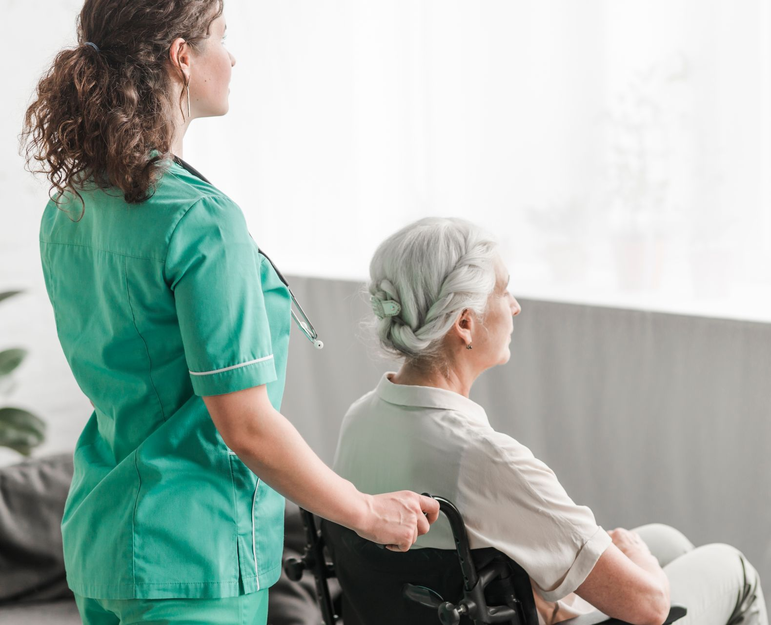 Nurse in green scrubs pushing a woman in a wheelchair, looking out a window.