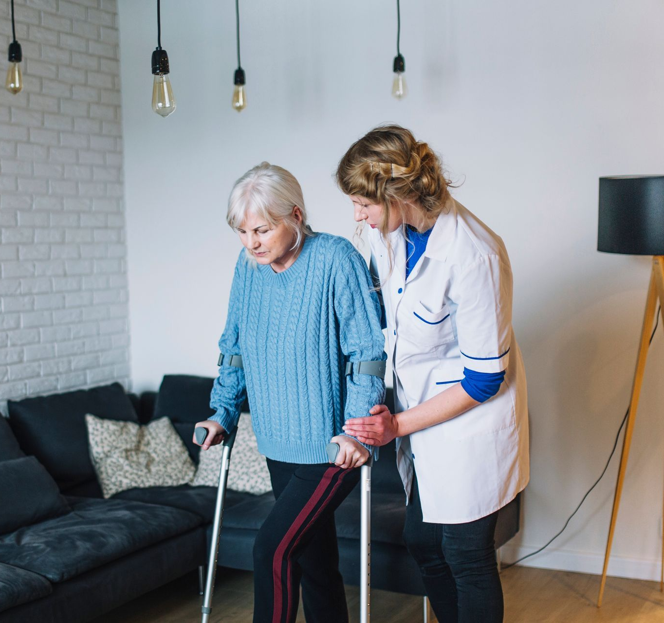 A woman with crutches is assisted by a caregiver while walking indoors.