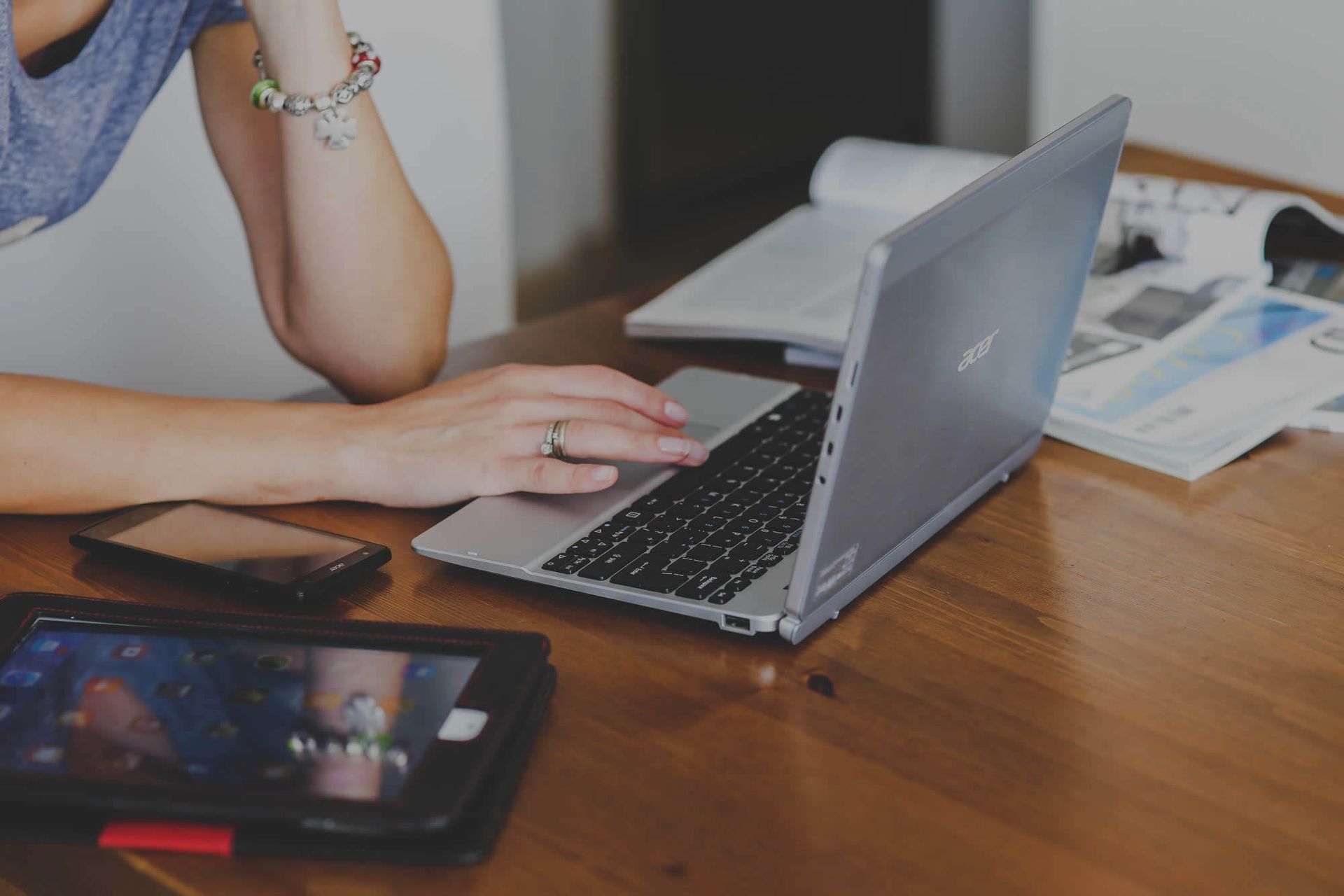 A person works on a silver laptop at a wooden desk with a tablet and papers nearby.
