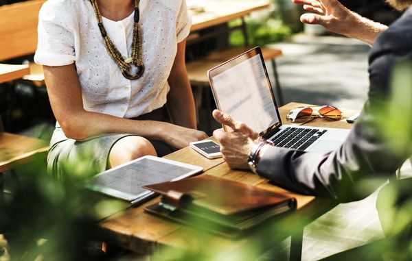 Two professionals sit at a wooden outdoor table, working with a laptop, tablet, and notebook during a meeting.