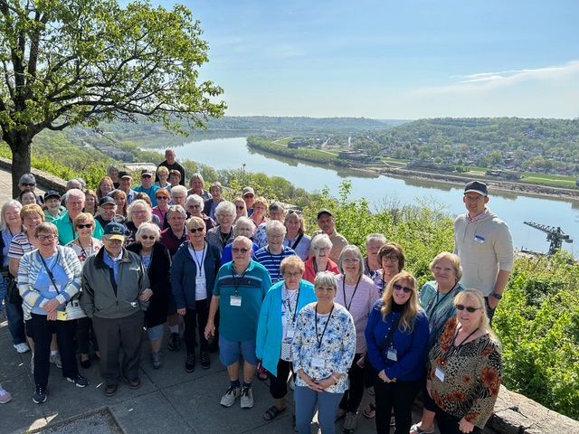 Group of people posing by a river. Sunny day, blue sky, trees, and buildings visible in the background.
