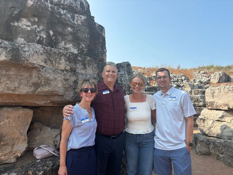 Four people pose together near ancient stone ruins, under a clear, sunny sky.