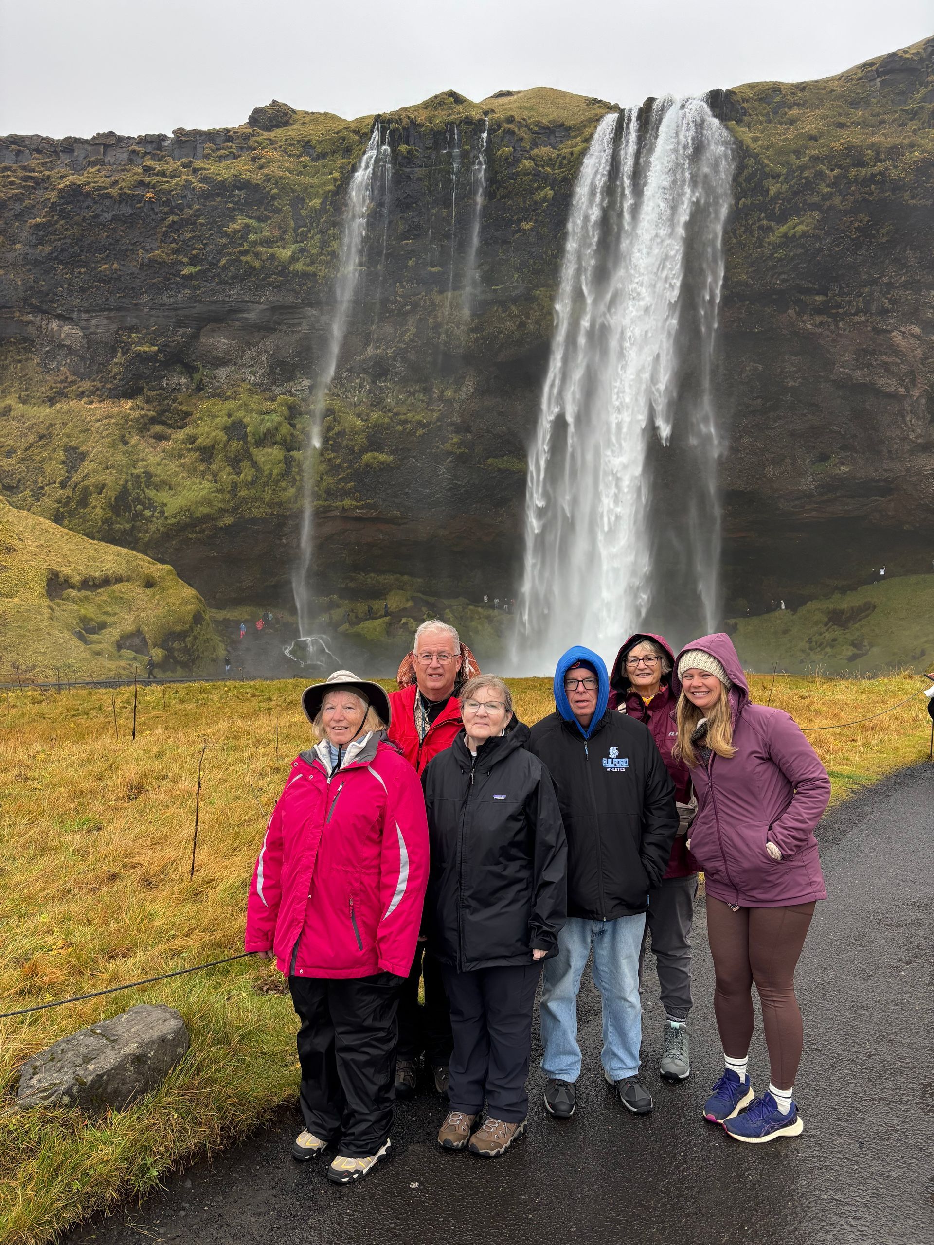 Group of people pose in front of Seljalandsfoss waterfall in Iceland. Cloudy sky, wearing jackets, and enjoying the scenery.