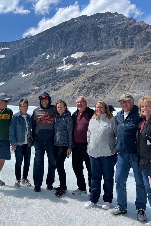 Group of people pose on ice with a mountain backdrop, wearing casual clothes on a sunny day.
