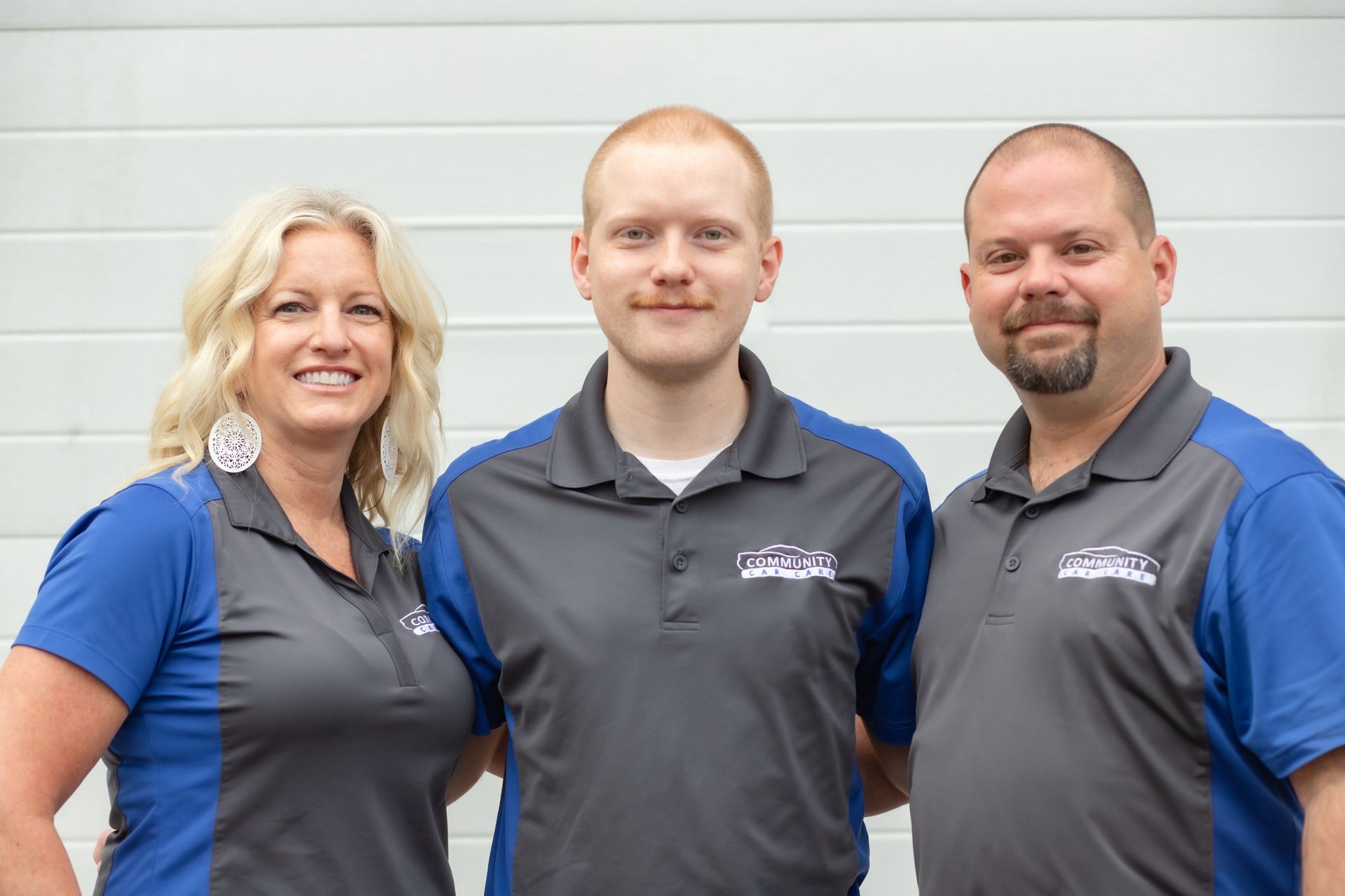 Three people in matching grey and blue polo shirts stand against a white slatted wall, smiling at the camera.