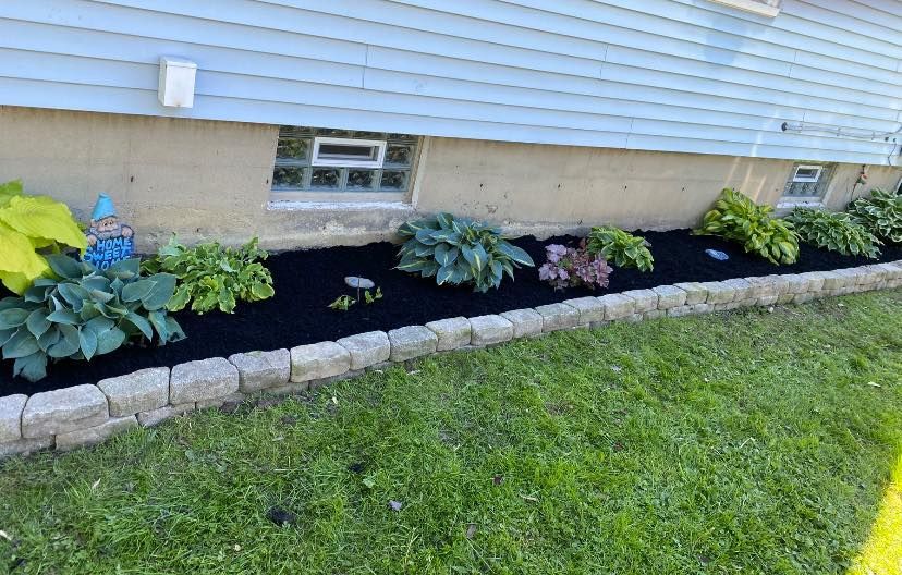 A flower bed with black mulch and green plants, bordered by stone blocks, beside a blue house.