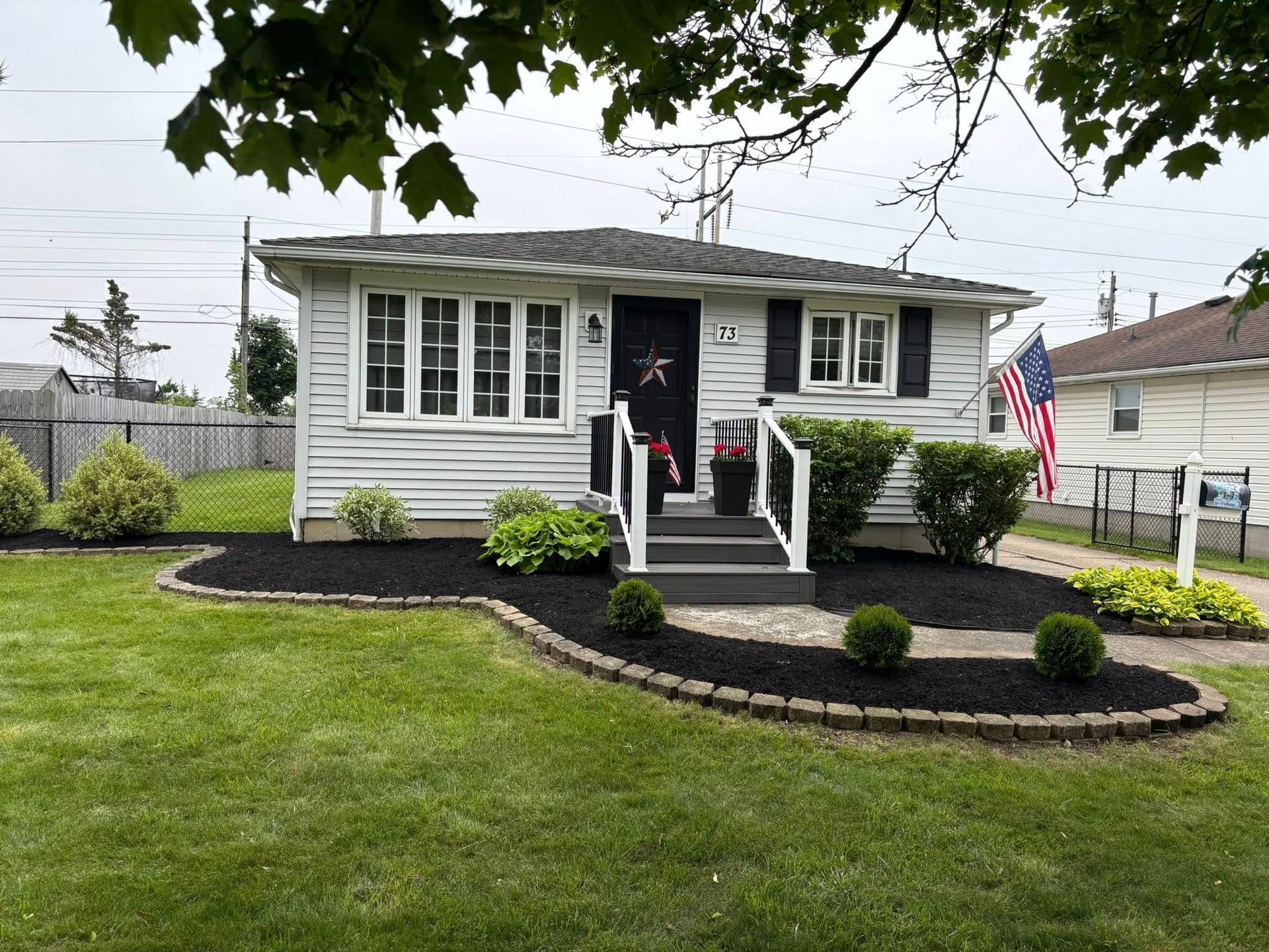 Small white house with black mulch landscaping, green lawn, American flag, cloudy sky.