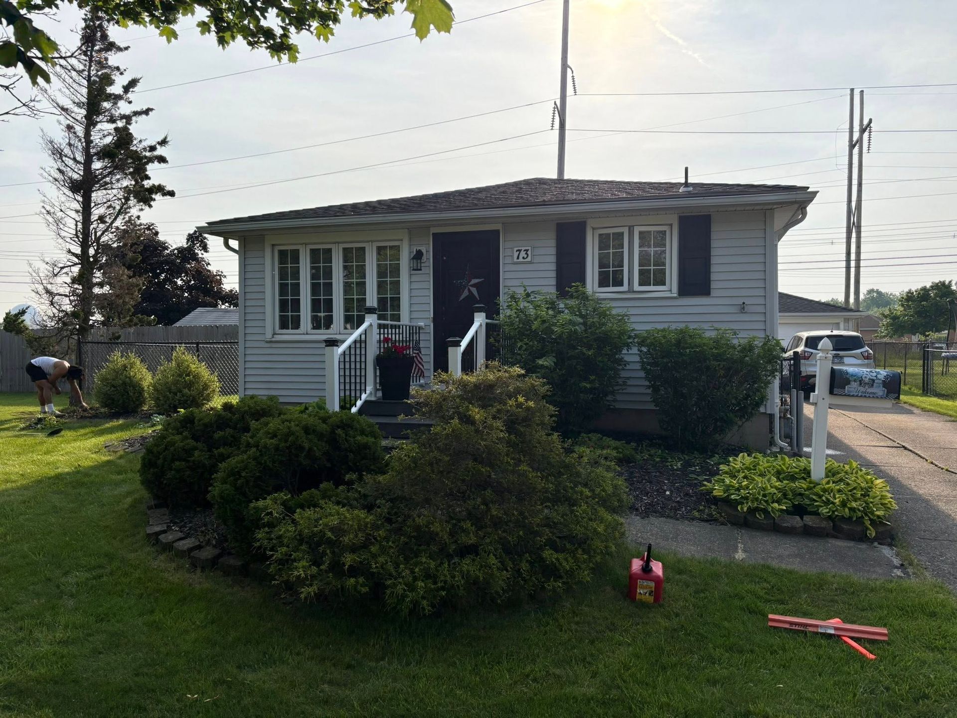 Small white house with bushes, front porch, and driveway on a sunny day. A person is working in the garden.