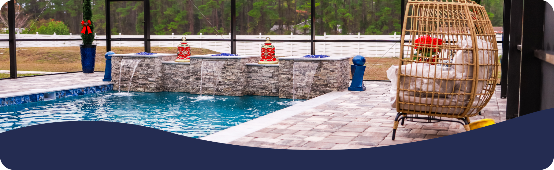 A pool with a stone wall fountain, decorative statues, and a rattan swing chair on a patio inside a screened enclosure.