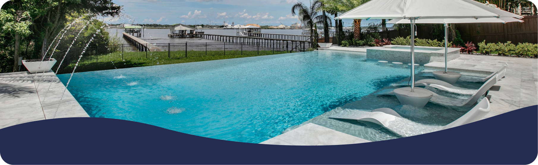 An infinity pool with lounge chairs and an umbrella, overlooking a calm waterfront with a dock under a bright blue sky.