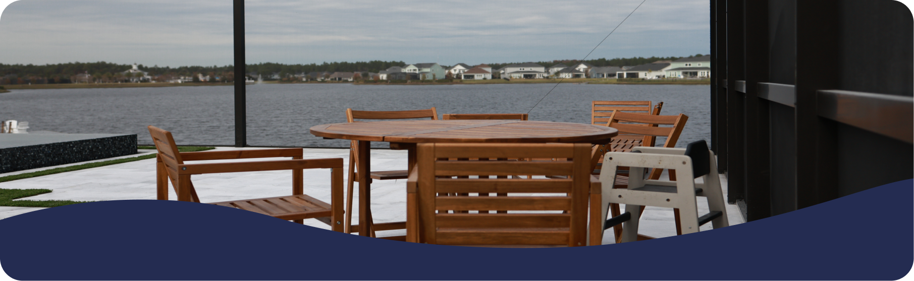 Wooden patio furniture sits on a deck overlooking a lake with a distant shoreline under a cloudy sky.