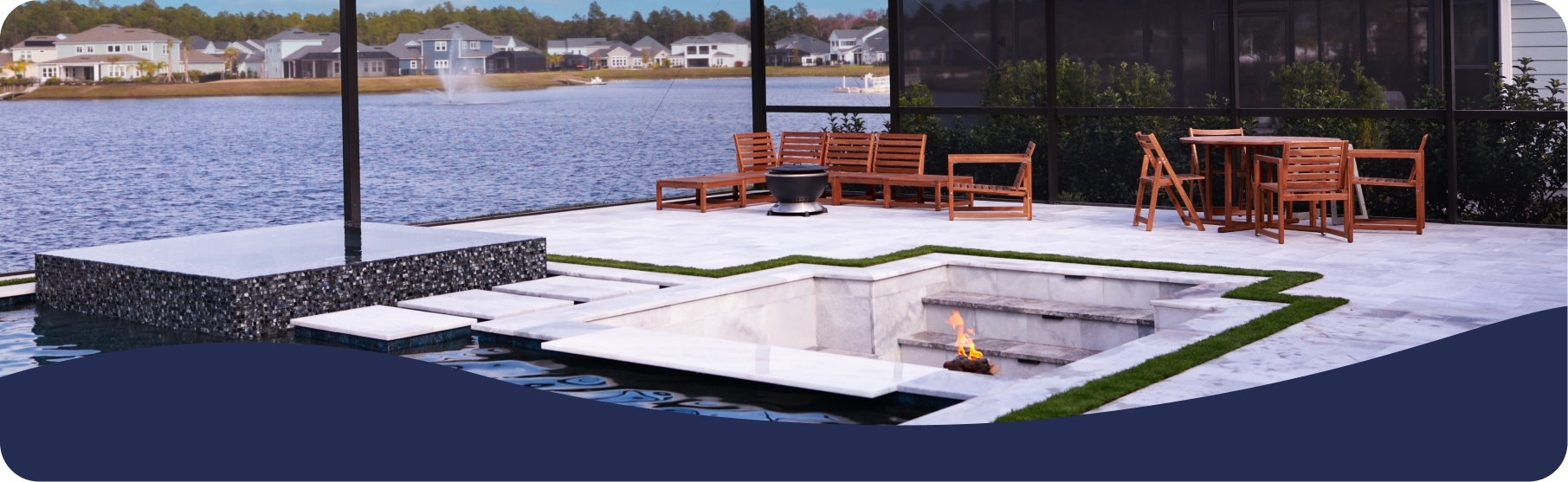 A patio beside a lake with a sunken fire pit, stone flooring, and outdoor wooden furniture enclosed in a screen.