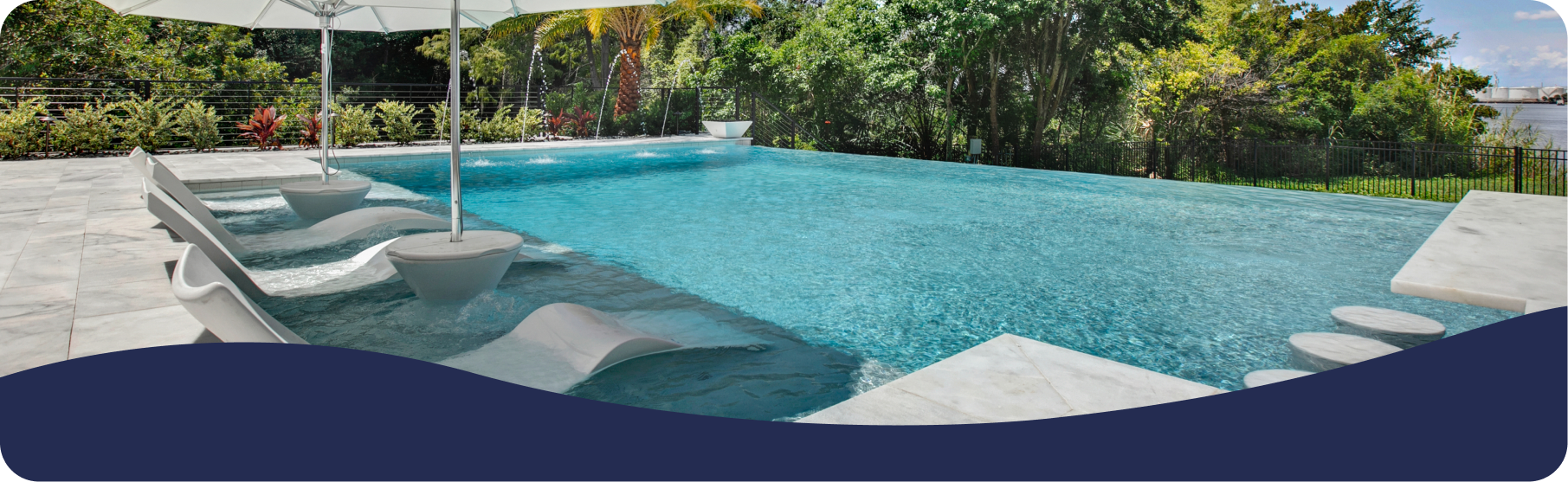 An infinity pool with submerged lounge chairs, a white umbrella, and a lush green background.