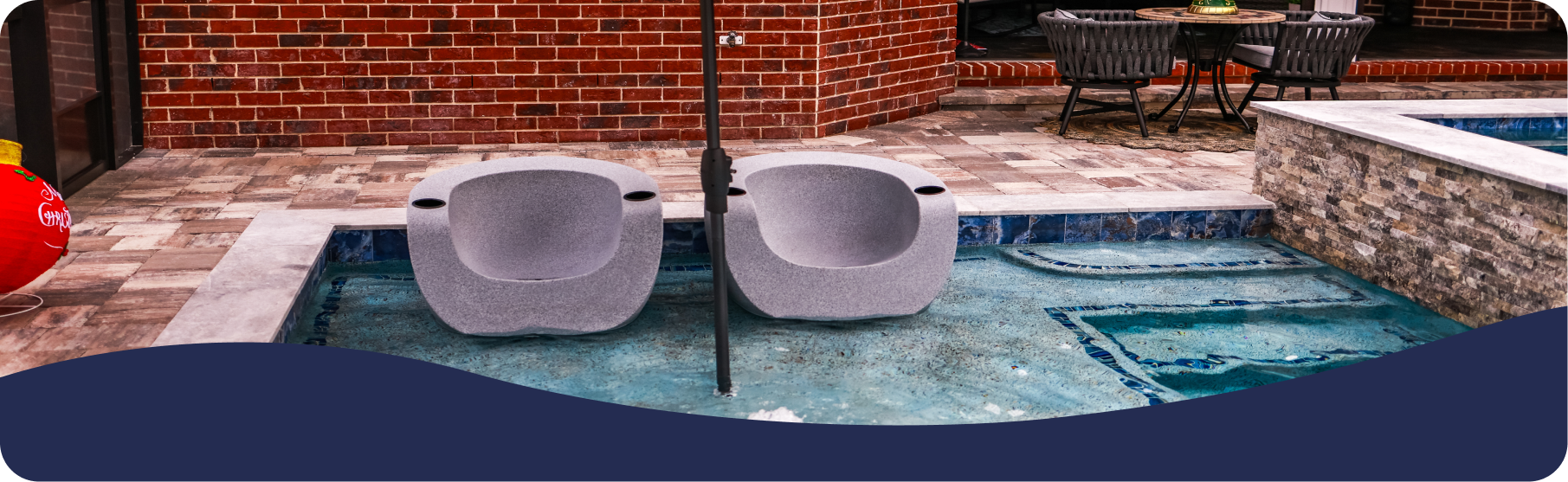 Two modern gray pool chairs sit in shallow water on a patio next to a brick wall and outdoor dining set.