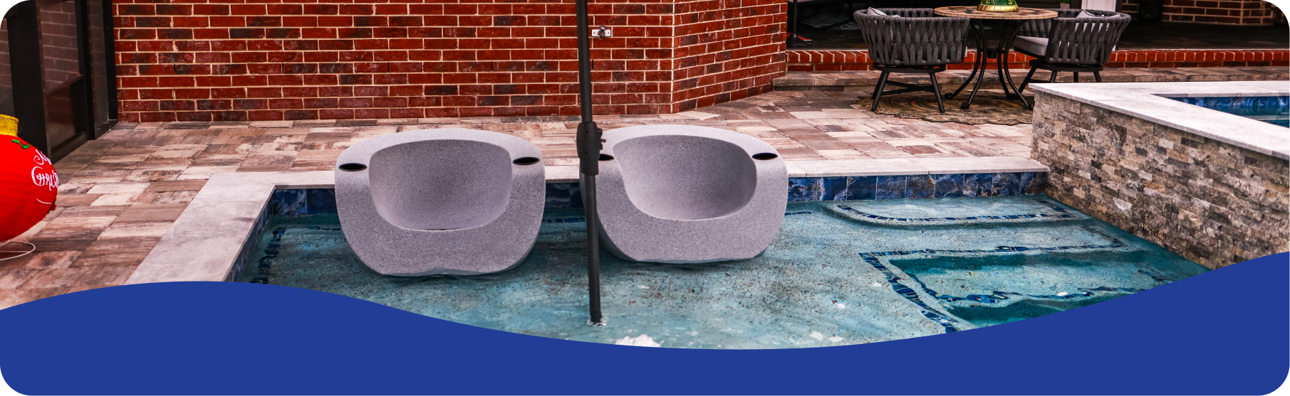 Two modern gray pool chairs sit in shallow water on a patio next to a brick wall and outdoor dining set.