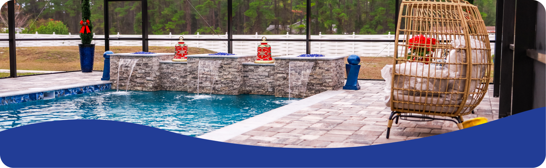 A pool with a stone wall fountain, decorative statues, and a rattan swing chair on a patio inside a screened enclosure.