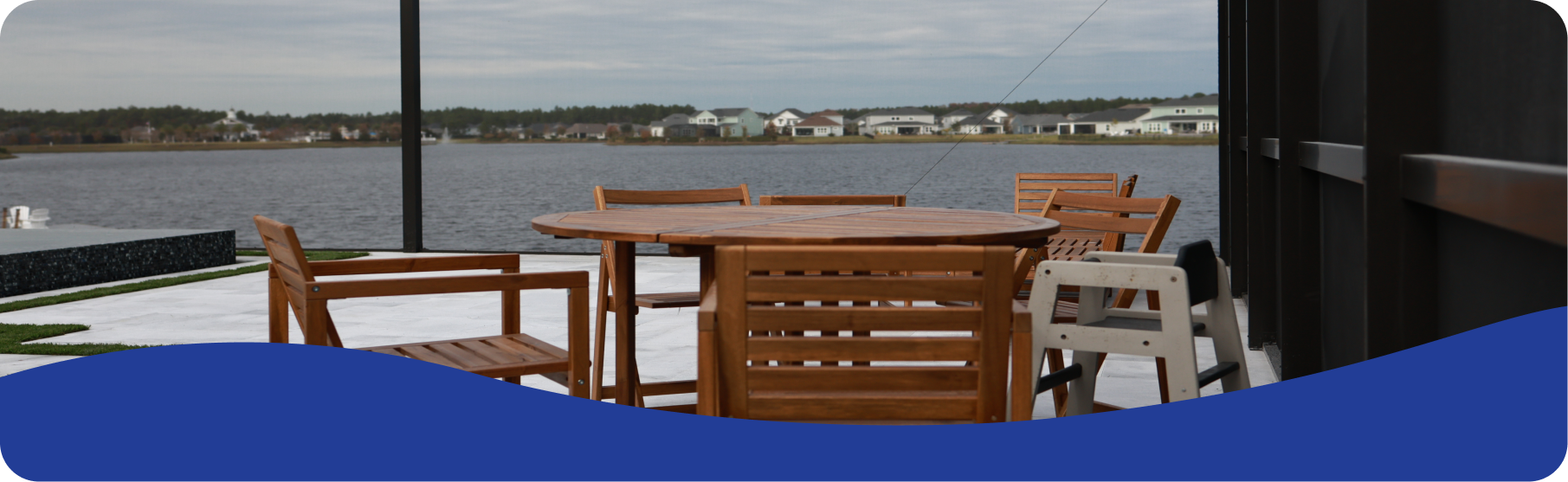 Wooden patio furniture sits on a deck overlooking a lake with a distant shoreline under a cloudy sky.