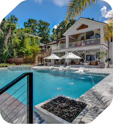 A swimming pool with white umbrellas and a fire feature in the foreground, set against a two-story house under blue skies.