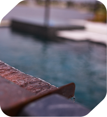 A close-up view of water trickling over a rustic, metal edge into a dark, calm pool.