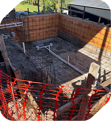 A swimming pool under construction showing rebar framing, white plumbing pipes, and orange safety fencing in an outdoor lot.