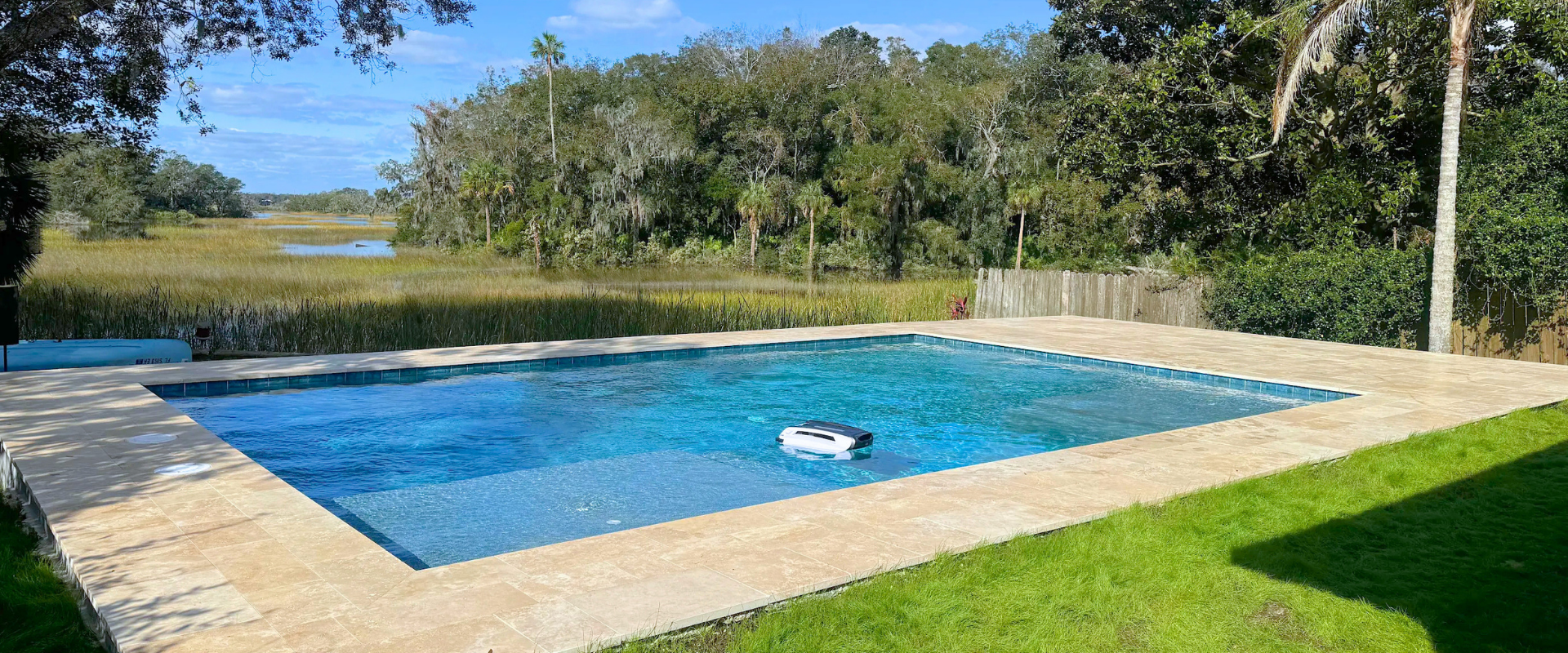 Blue swimming pool with a floating cleaner, surrounded by a stone deck and green lawn under trees