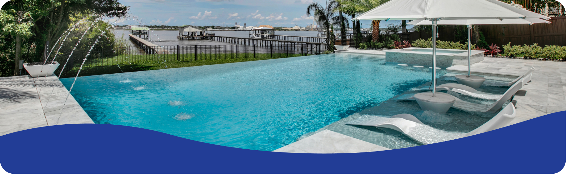 An infinity pool with lounge chairs and an umbrella, overlooking a calm waterfront with a dock under a bright blue sky.