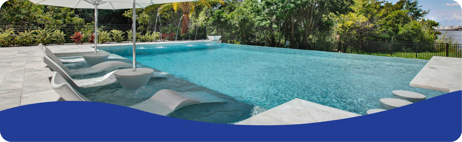 An infinity pool with submerged lounge chairs, a white umbrella, and a lush green background.