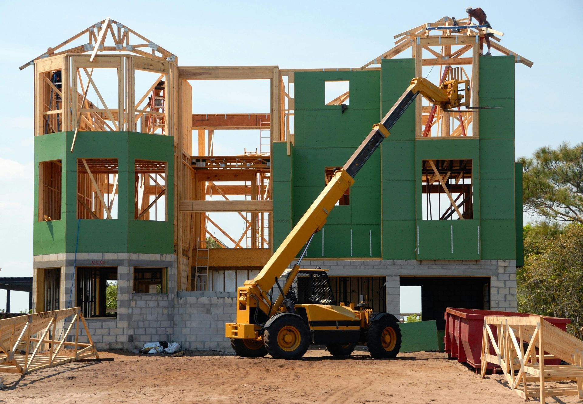 Construction of a two-story house frame, with a yellow telehandler crane. Green sheeting covers exterior.