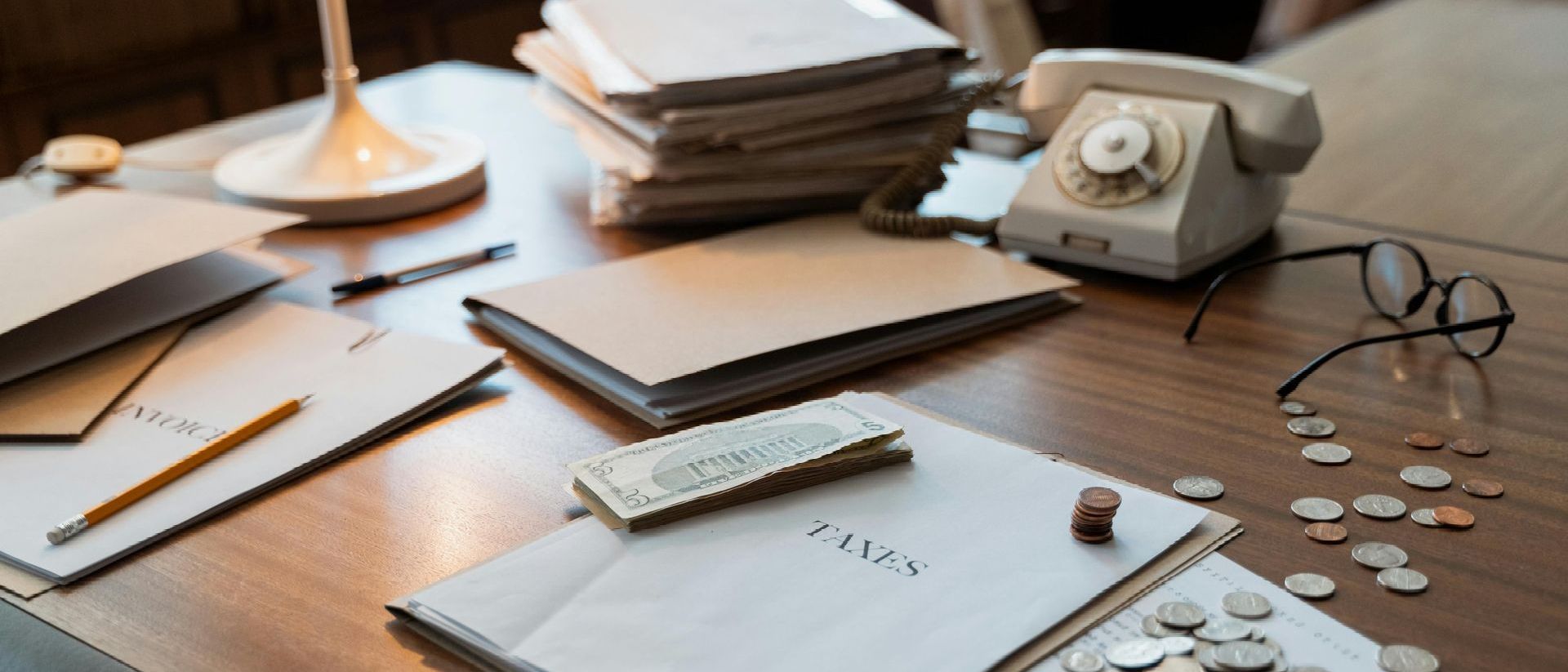 A desk with a stack of papers , a telephone , coins , and glasses.