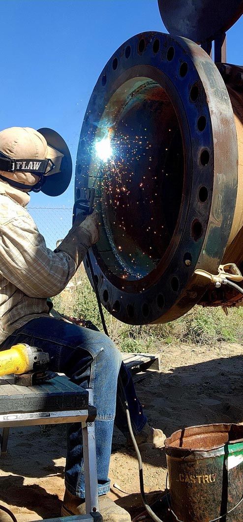 A man is welding a large piece of metal while sitting on a bench.