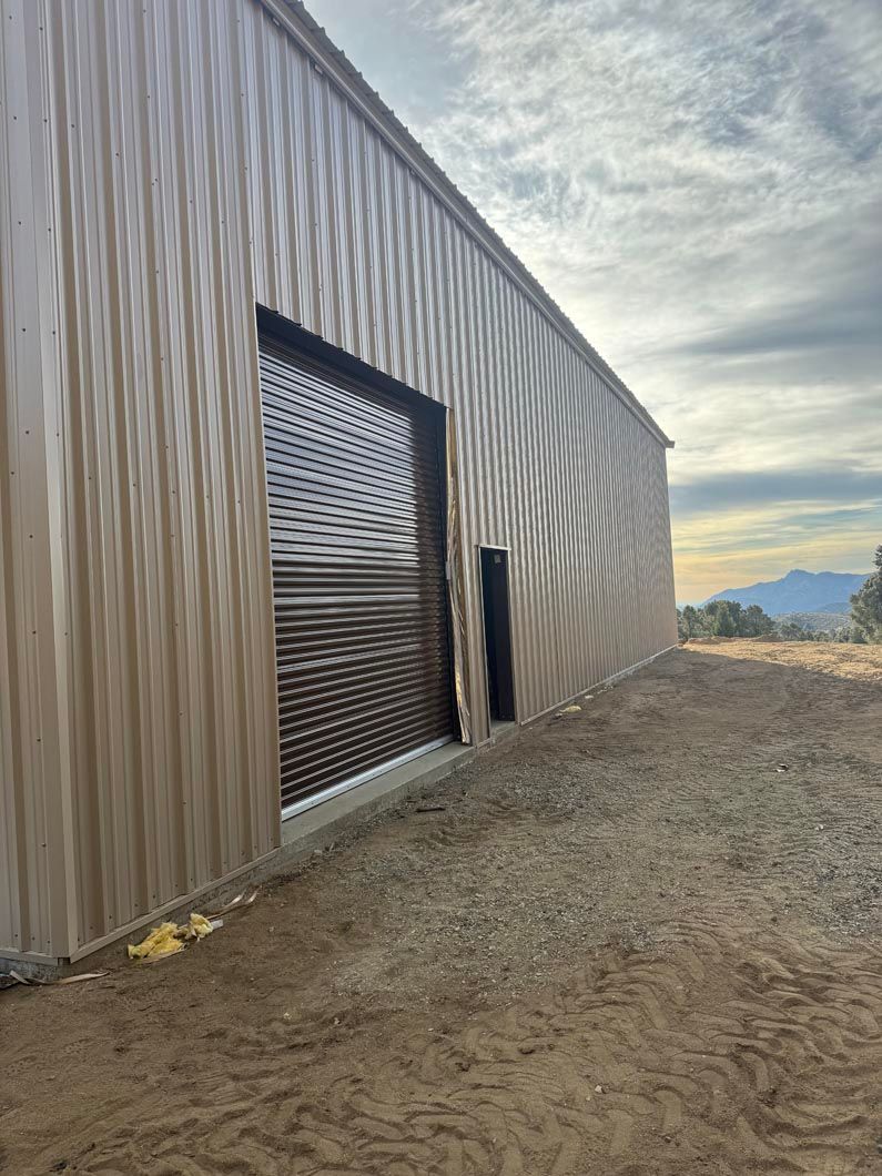A large metal building with a garage door is sitting in the middle of a dirt field.
