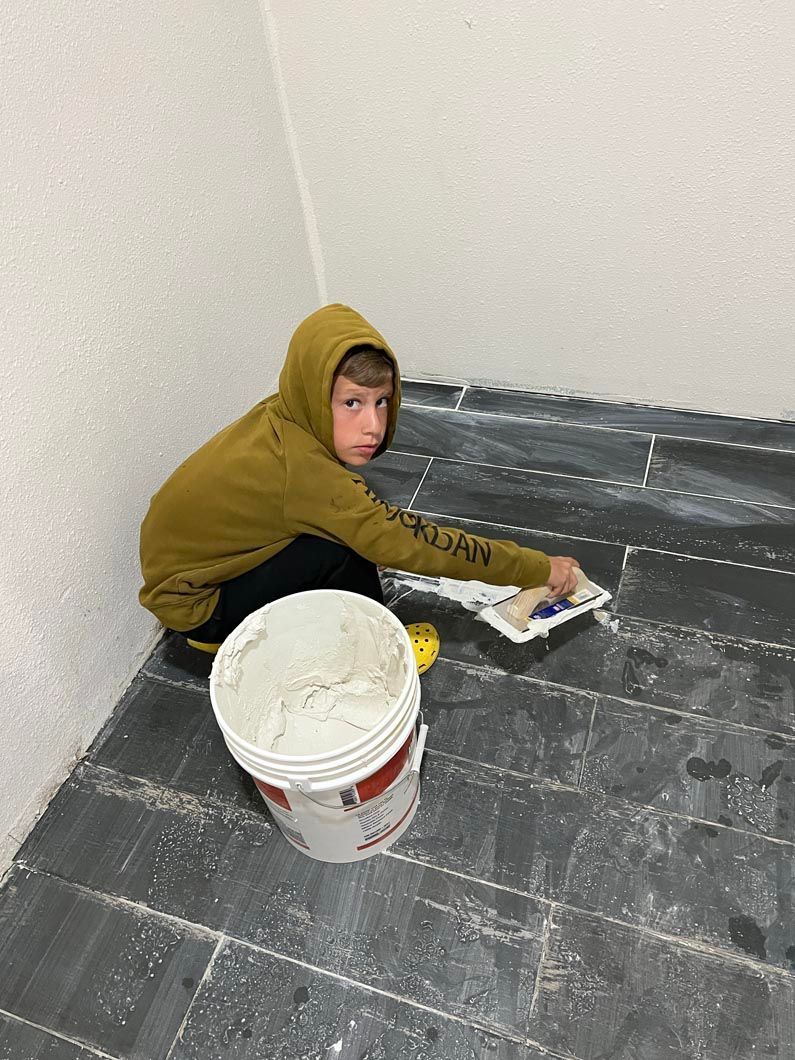 A young boy is sitting on the floor next to a bucket of cement.