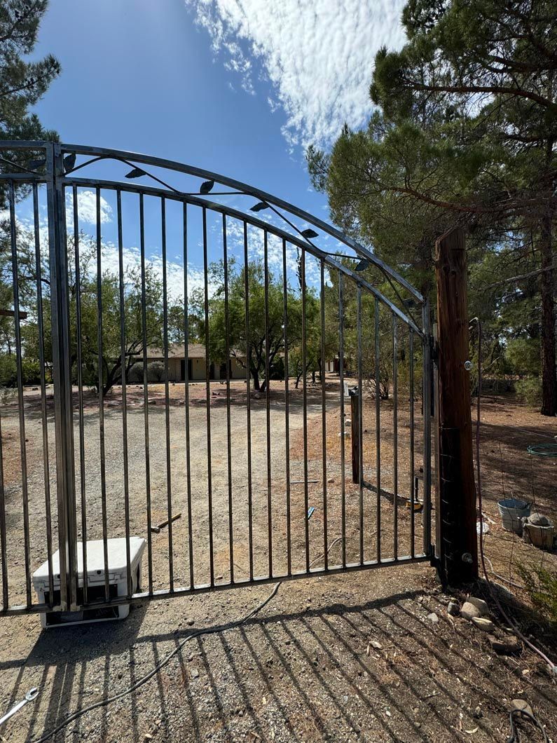 A metal gate is open to a dirt field with trees in the background.