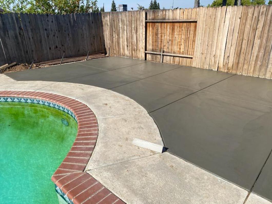 A concrete patio next to a swimming pool with a wooden fence in the background.