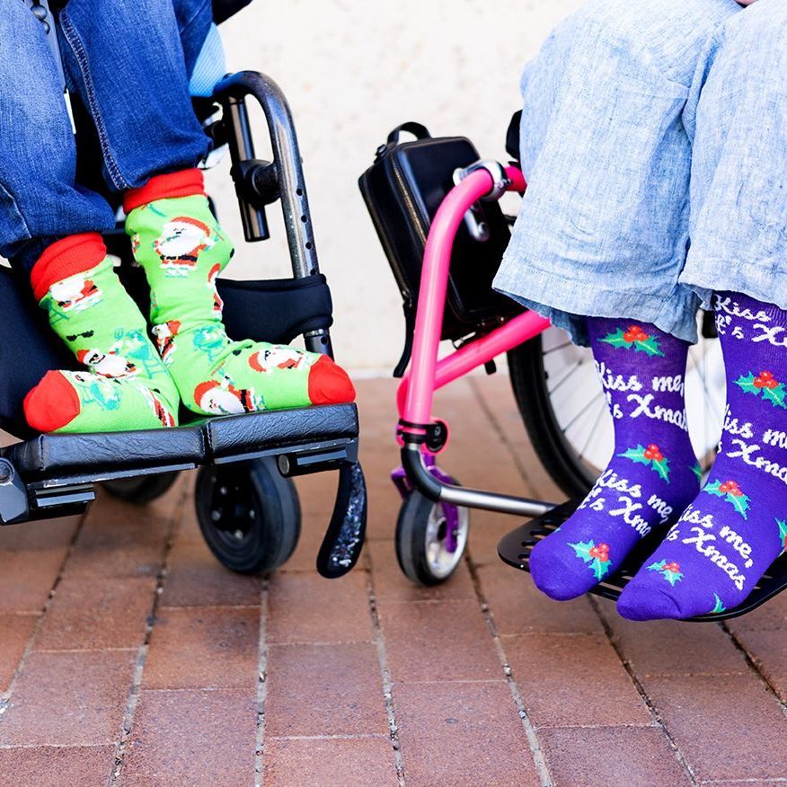 A close-up of two wheelchair users’ feet, both wearing Christmas themed
socks. The individual on the left is wearing green Santa socks and the individual on
the right is wearing purple socks that say ‘kiss me it’s X-mas’.