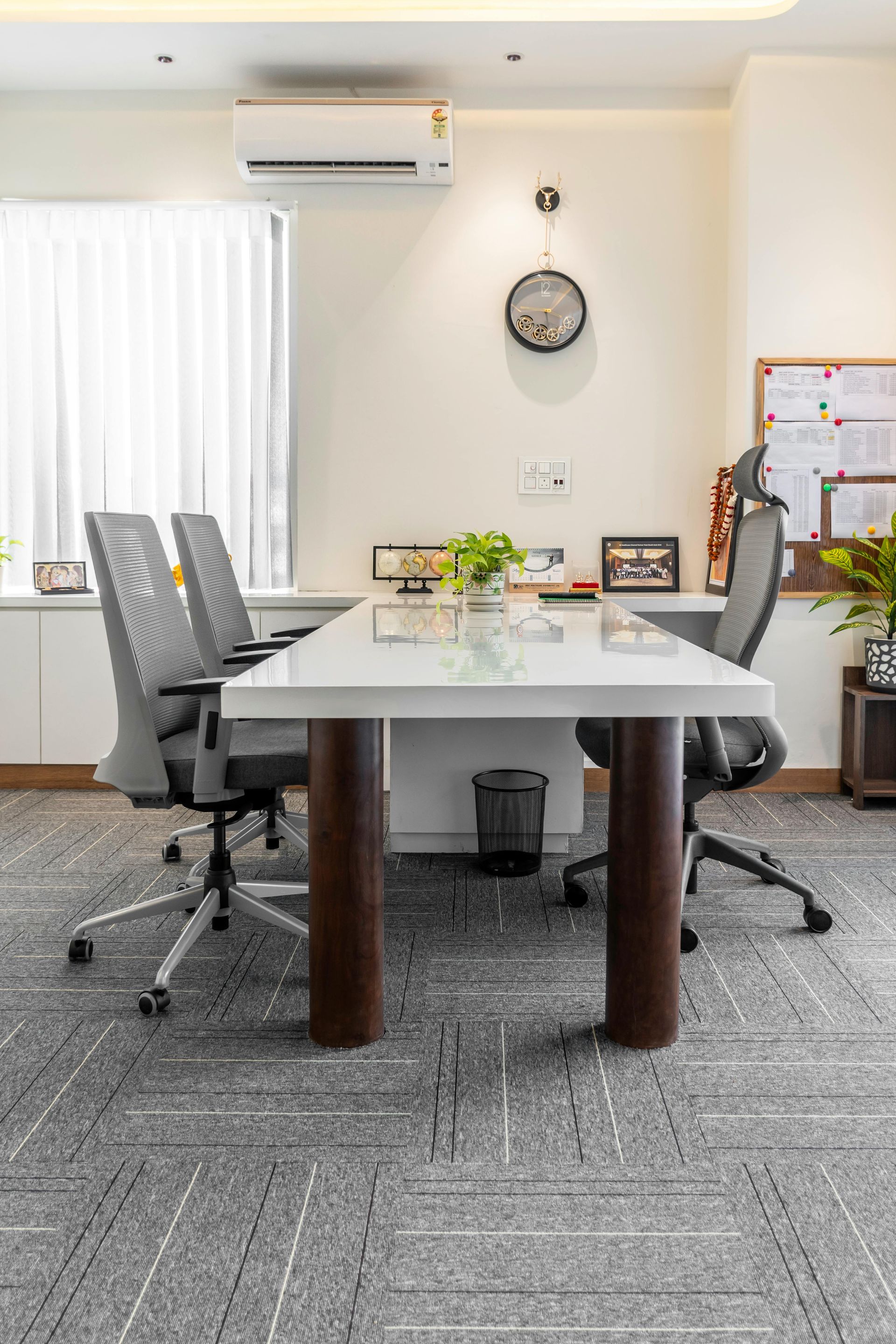 Office with a white table, gray chairs, and gray carpet; a window, clock, and bulletin board are on the wall.