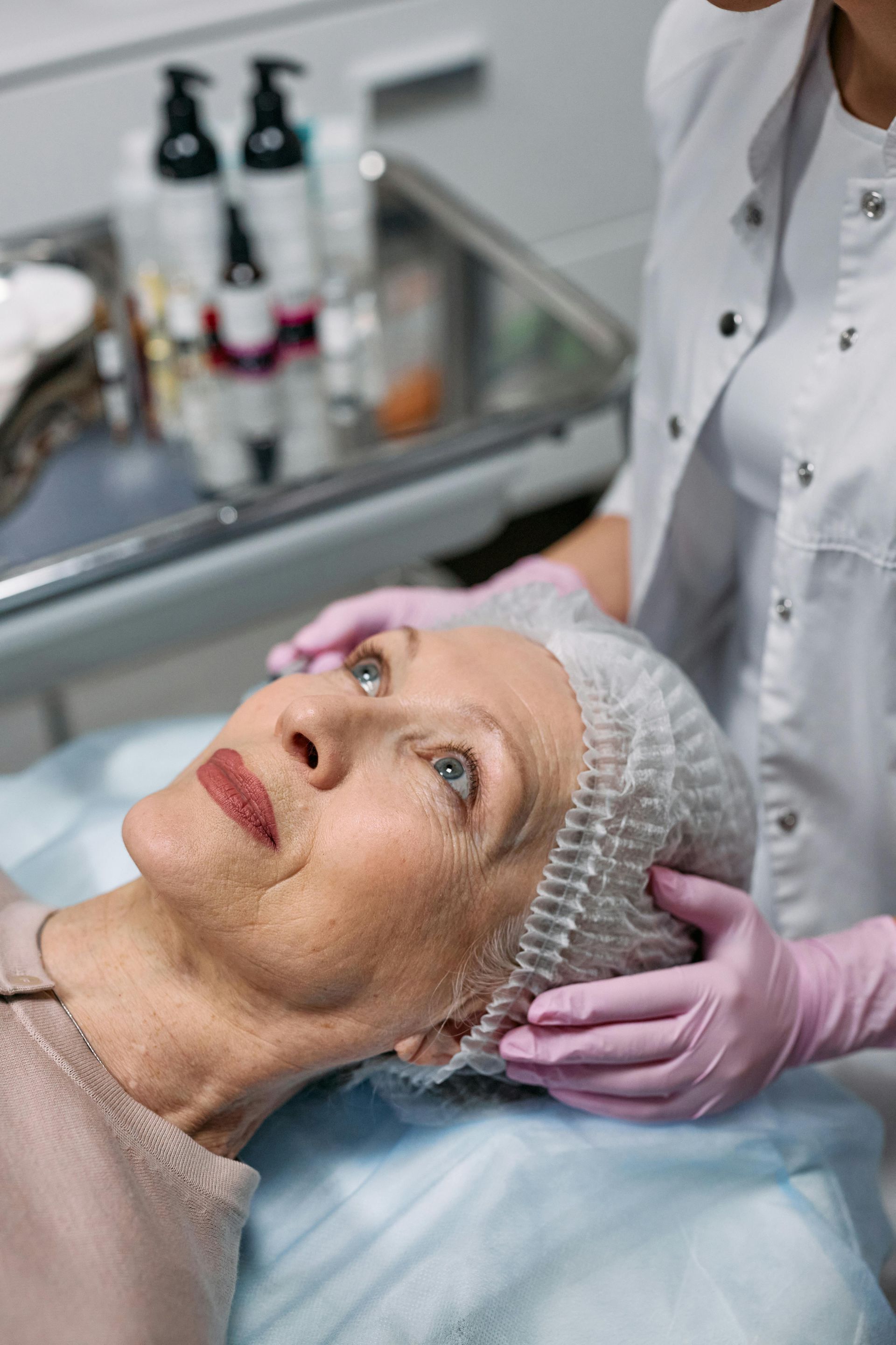 Woman receiving facial treatment in a clinic. Person in gloves examines her face. Products visible in background.