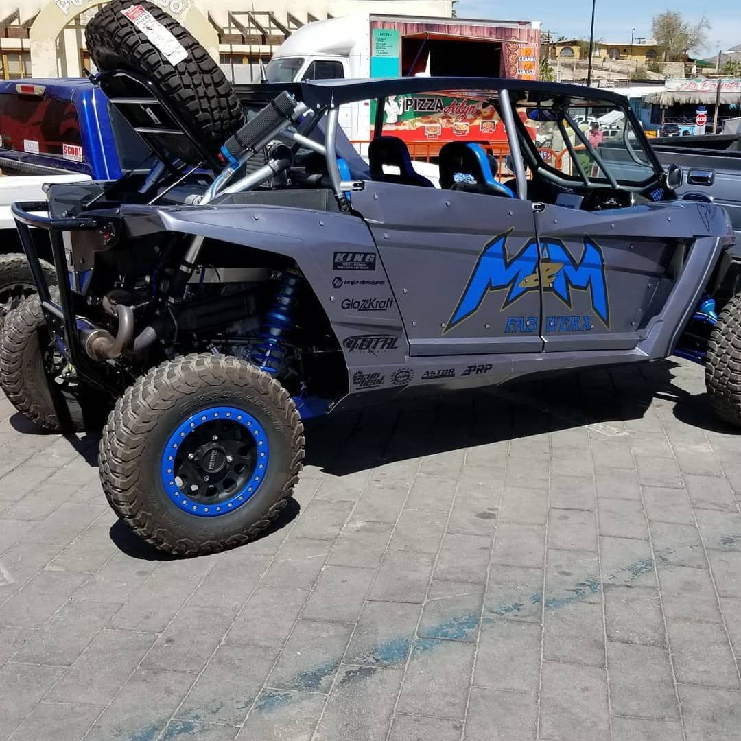 Gray off-road dune buggy with blue accents and oversized tires parked outdoors near other vehicles.