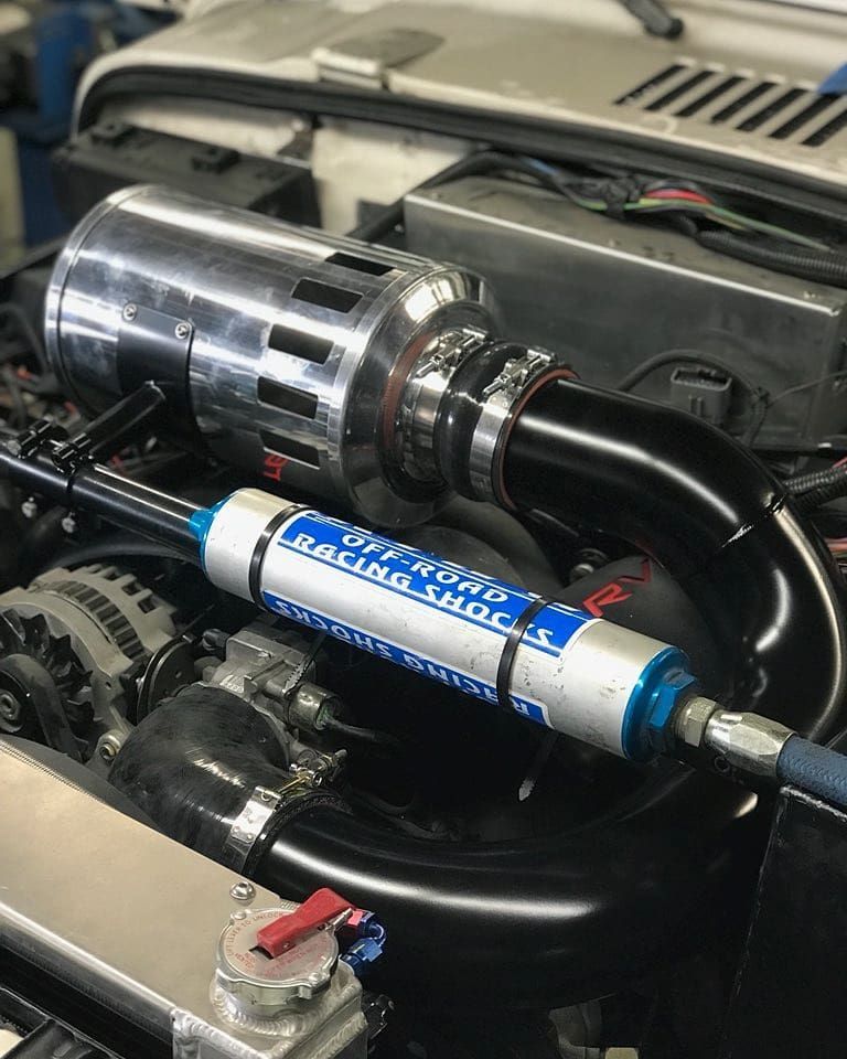Car engine bay with polished metal intake and blue cylindrical aftermarket component connected by black hoses